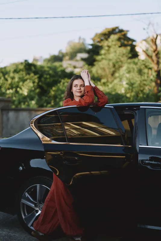 A woman in a red dress standing beside a sleek car on a sunny street, capturing modern lifestyle and fashion in an outdoor urban setting. Natural light highlights confidence and elegance