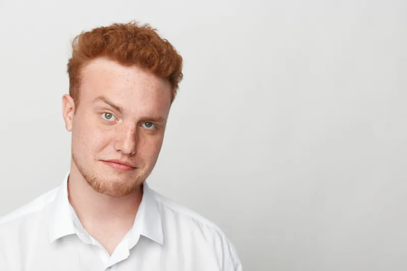Portrait of a young redhead man with freckles wearing a white shirt looking at camera. Studio shot on white background with natural expression and casual style. Concept of youth and confidence.