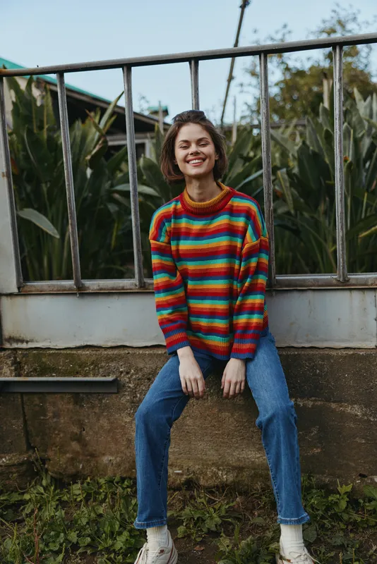 Woman in a colorful striped sweater smiling while sitting on a low wall, casual jeans and sneakers, relaxed outdoor portrait with natural plants and metal railing background.