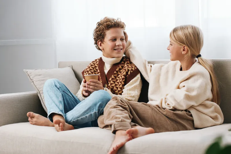 Authentic scene of two children at home, sharing a cozy moment on a sofa, wearing warm sweaters, enjoying playful time and a simple snack, in a peaceful afternoon setting.