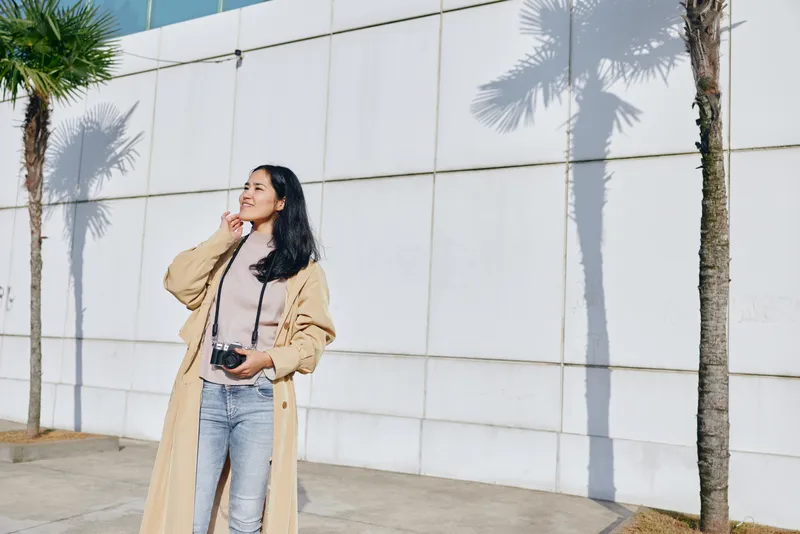 Woman with camera stands outdoors beside a light wall, wearing a beige coat and jeans, enjoying sunlight, shadows, and palm trees in a bright urban setting.