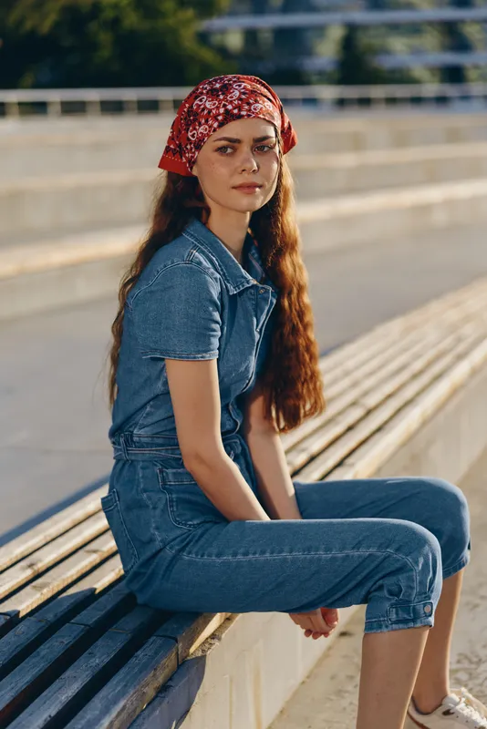 Modern boho-western denim street style woman with long hair and red bandana sitting outdoors, natural light, film color tones, casual lifestyle fashion portrait.