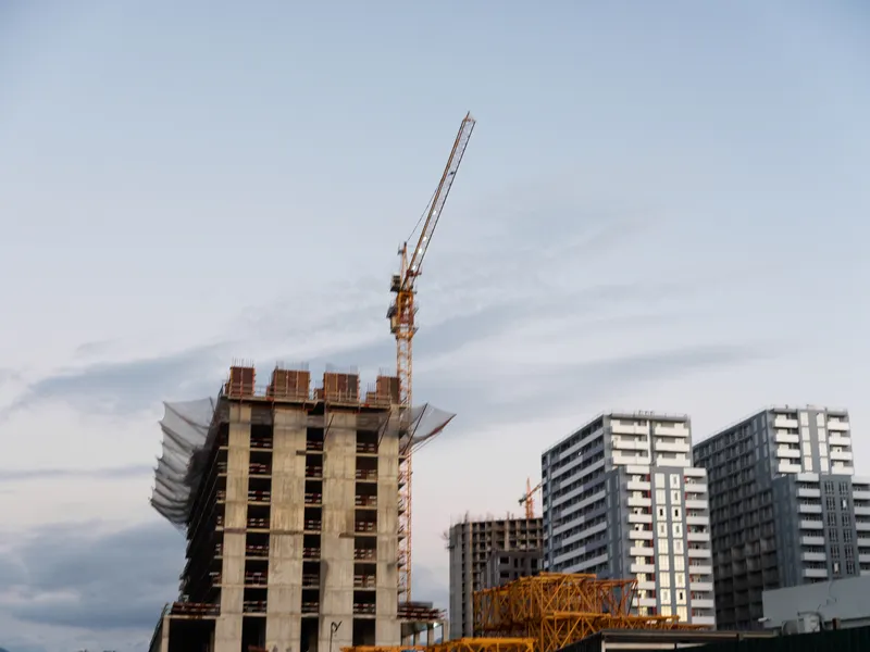 A construction site features a tall concrete building under development with a towering crane, scaffolding, and heavy equipment, creating an active urban project scene.