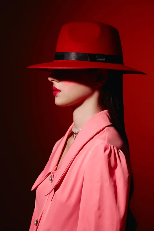 Avant-garde woman in a red fedora hat and pink trench coat stands in profile against a red background. Concept of bold beauty, fashion, and creative editorial style.