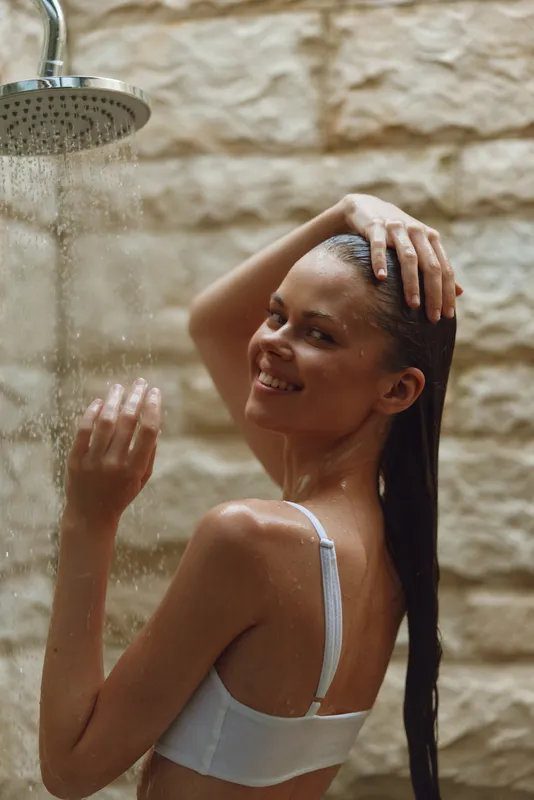 smiling woman enjoying shower with water droplets, long wet hair, natural skin tone, relaxed atmosphere, beige stone background, intimate moment