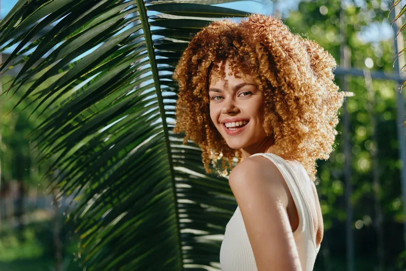 smiling woman with curly hair outdoors wearing white tank top near tropical palm leaves in sunny natural environment