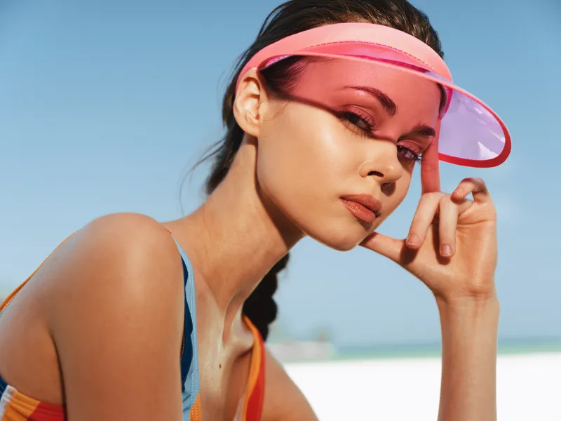 Close up portrait of a sunlit woman on a beach, wearing a pink visor and colorful swimsuit, with a calm expression against blue sky and white sand.
