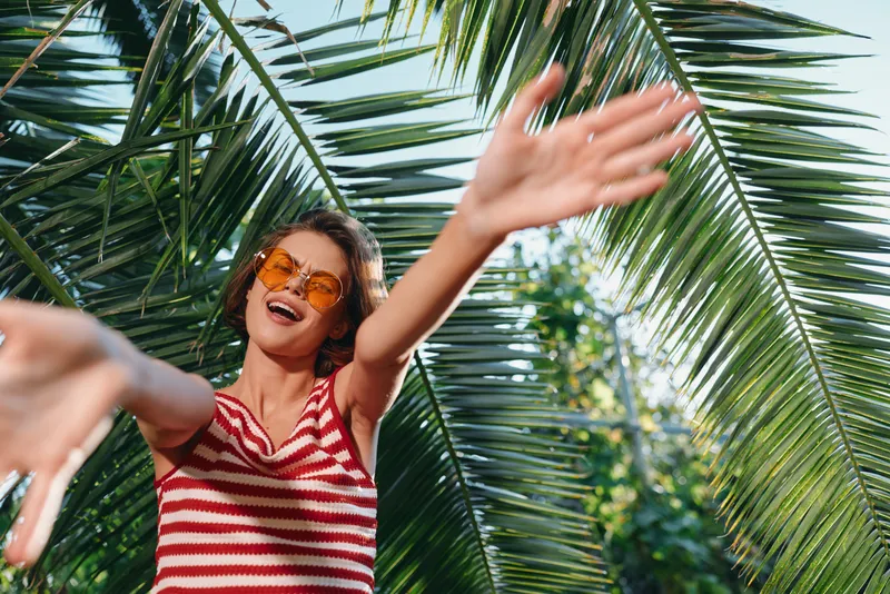 A cheerful woman wearing a striped sleeveless top and sunglasses waves joyfully under palm fronds, capturing a bright, carefree moment in a tropical outdoor setting.
