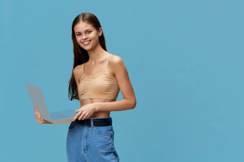 Young smiling woman holding laptop while standing against blue background, wearing casual beige top and jeans, conveying productivity and modern lifestyle.