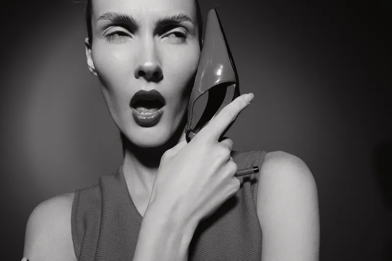 Closeup of a white woman with light skin holding a high heel shoe to her face in a dramatic black and white portrait. Concept of fashion and expression.