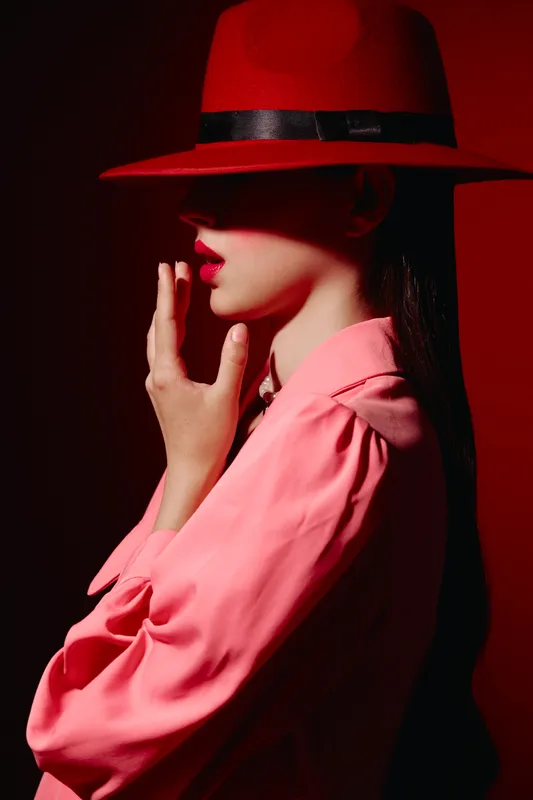 Woman in a red hat and bright clothing poses with her hand near mouth. Studio portrait with high contrast and dramatic shadow on a dark background. Fashion expression concept.