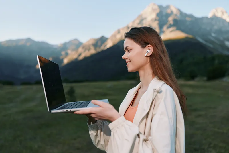 A cheerful professional woman stands outdoors in a scenic mountain setting, holding a laptop and smiling as she types, conveying focus, mobility, and a modern work lifestyle.