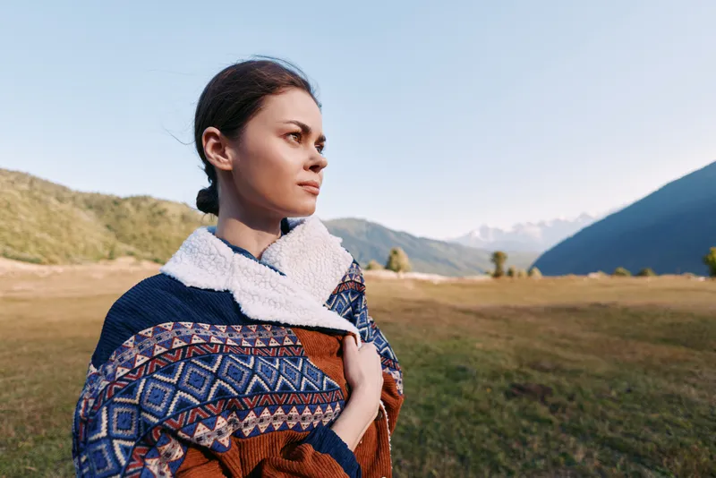 woman portrait mountains landscape shawl nature outdoors traveler standing in grassy valley looking into distance with calm gaze and contemplative mood, warm light on scenic countryside scene