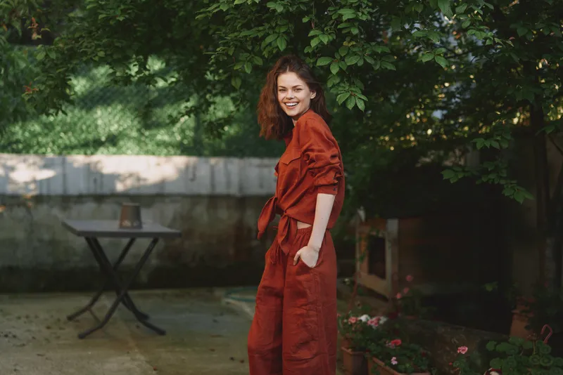 Young woman wearing rust overalls smiles softly in a green garden courtyard, creating a relaxed, confident lifestyle moment