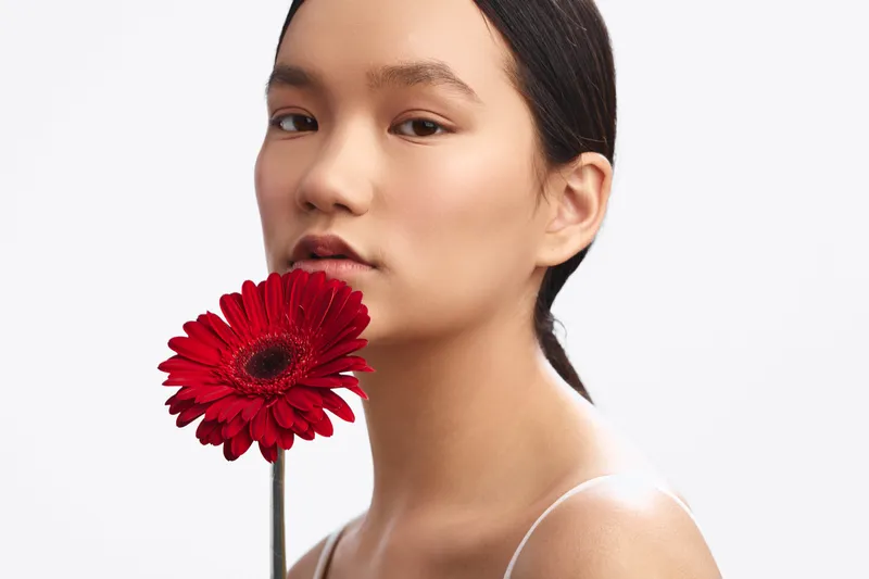 asian woman holds a bold red flower near her face in a minimal studio. her calm expression and soft lighting create an intimate, artistic portrait with a serene, focused mood.