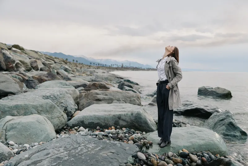 Woman standing on rocky shore wearing coat looks up with closed eyes by calm sea under cloudy sky. Concept of relaxation, freedom, nature, and peaceful moment outdoors.