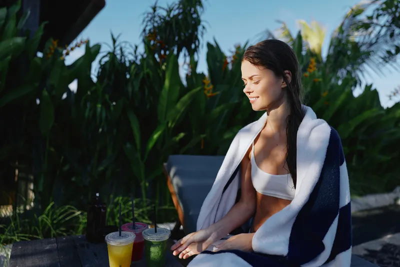 A young woman wrapped in a towel sits outdoors in sunlight near tropical plants with two colorful drinks on a table. Concept for relaxation, lifestyle, and healthy living.