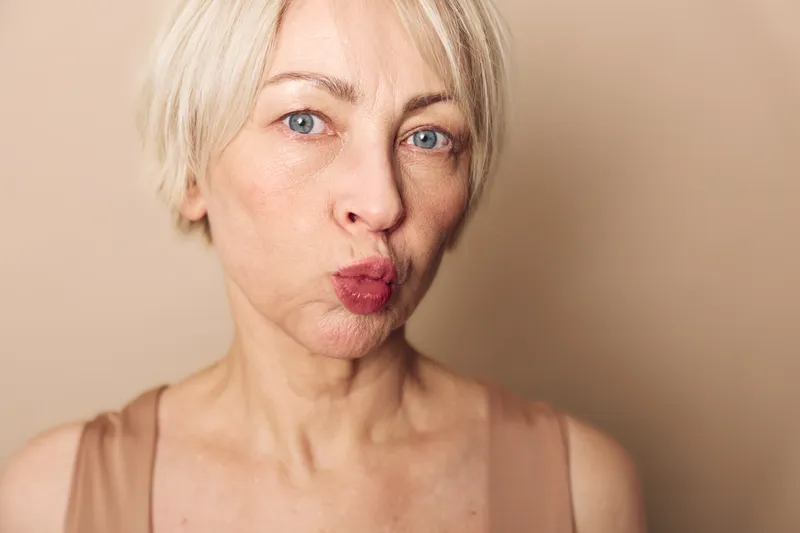 Closeup of mature woman with short blonde hair making a playful kiss face expression against beige background. Concept for natural beauty, aging, and confident female emotions.