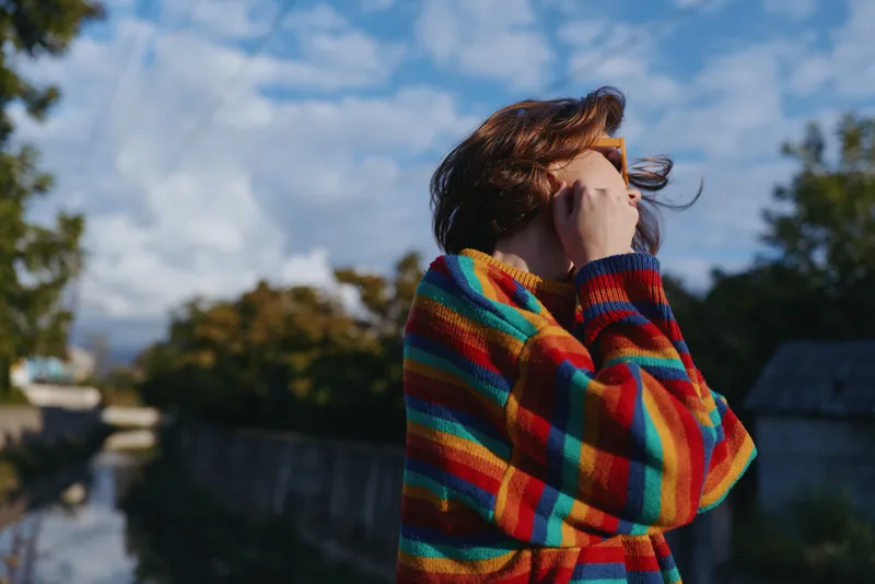 Woman in a rainbow sweater with short hair outdoors by a canal, contemplative and touching her face in autumn light, casual outfit, candid travel lifestyle portrait.