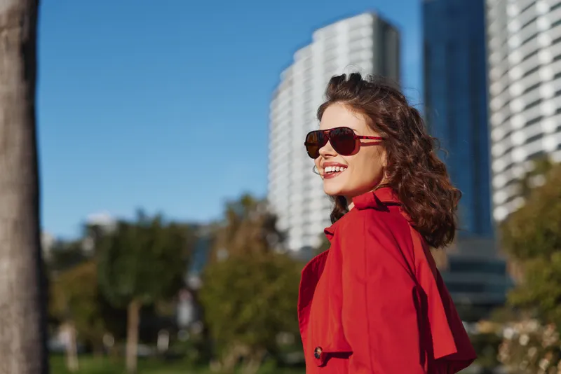 Smiling woman with curly hair wearing sunglasses and red coat outdoors in urban park on sunny day. Modern buildings and green trees in background create lively atmosphere.