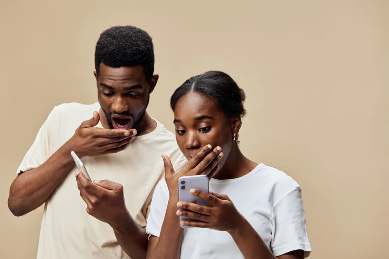 Surprised couple reacting to news on smartphones, showcasing diverse ethnicity and casual attire, against a light beige background