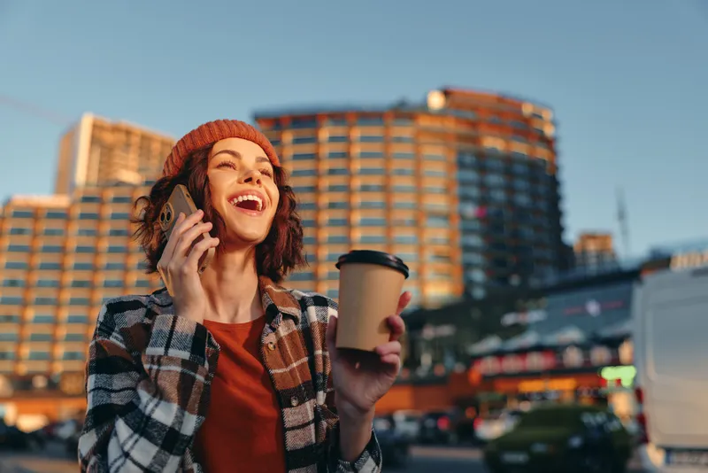 Beaming woman holds coffee and phone in city street, smiling in urban morning with golden hour glow; candid lifestyle, authenticity and emotional storytelling convey mindful living and joy.