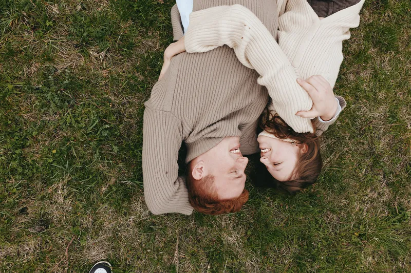 A smiling couple lies on grass in matching sweaters, facing each other and embracing in a bright outdoor setting. A genuine lifestyle moment of connection and warmth, illuminated by natural light.
