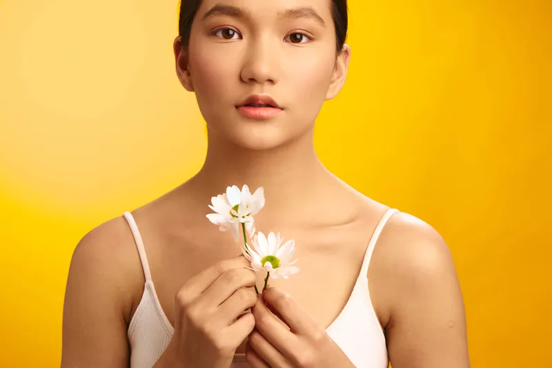 Asian woman standing against a vibrant yellow backdrop, gently holding white flowers close to her chest, radiating calm confidence, grace, and a fresh natural vibe.
