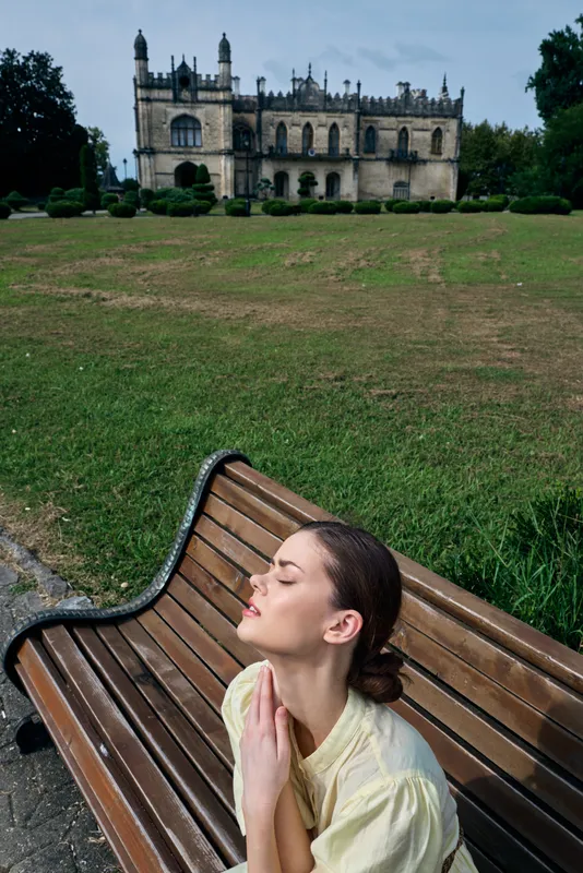 A woman sits on a bench in an outdoor setting, hands in prayer near her chest, with a grand castle in the distance creating a serene, contemplative moment and tranquil heritage.