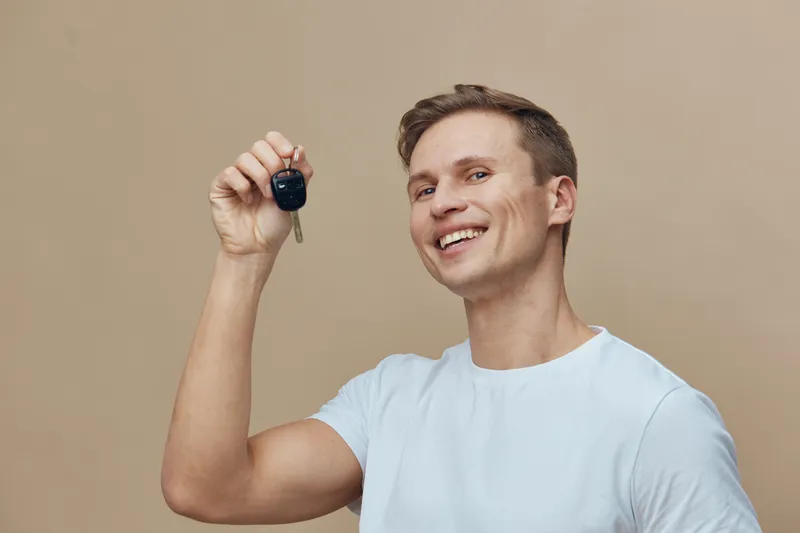 Smiling young man holding car key in hand, casual white t-shirt, positive facial expression, indoor studio shot with plain beige background. Happy lifestyle concept