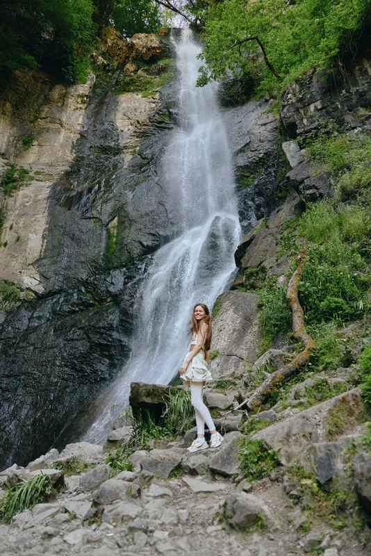 woman standing near waterfall in nature with rocky terrain and green trees wearing white outfit outdoors in daylight peaceful atmosphere