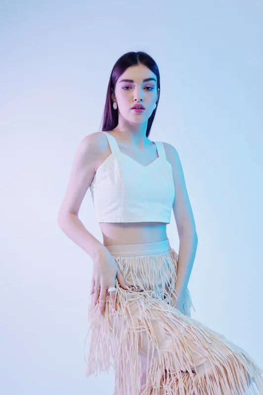 Young woman beauty fashion model poses confidently in a white crop top and fringed skirt. Studio shot with soft blue lighting. Concept for style, elegance, and modern trends.