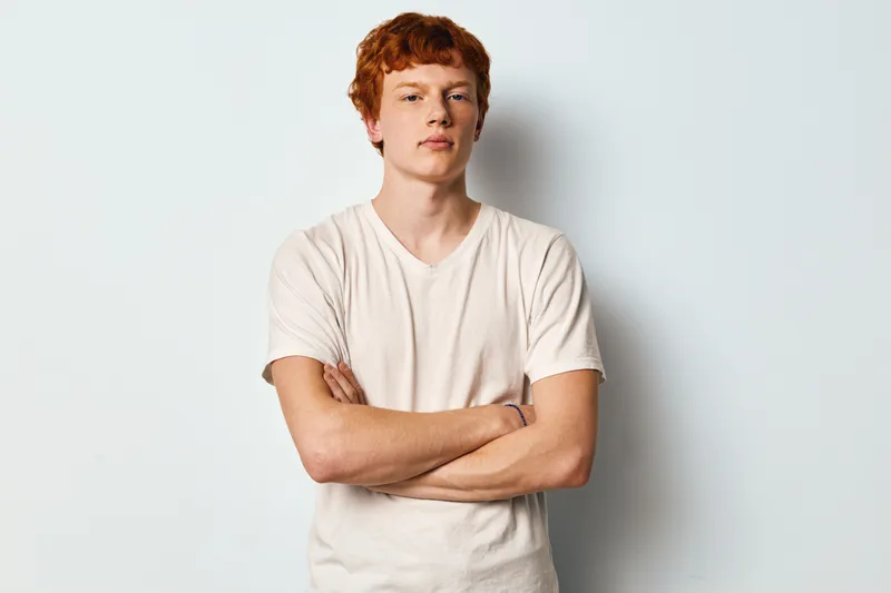 Young man with red hair and freckles stands with crossed arms wearing white t shirt on light background. Male portrait with confident expression and casual clothing in studio.