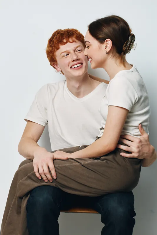 Young man and woman smiling and sitting close together on chair, casual clothing and happy expression with affectionate and warm connection between them.