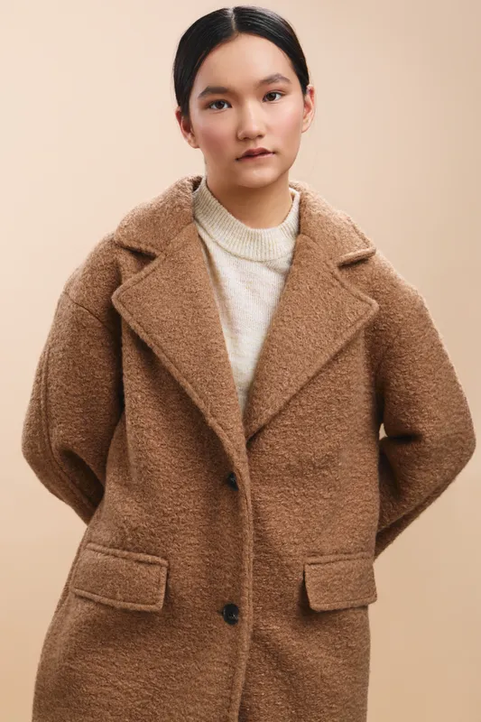 Asian woman stands in a neutral studio wearing a warm knit sweater and an oversized brown coat, projecting a calm, stylish mood and poised confidence.