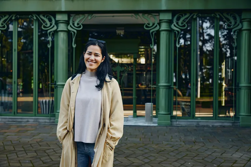 Woman in a beige trench coat smiles softly outdoors in front of a cafe with green ornate pillars, gray sweater and jeans creating a casual street fashion mood