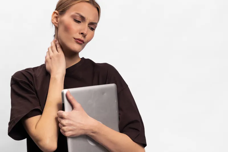 Young woman in dark shirt holds laptop and touches neck, looking worried. White background highlights her thoughtful mood, concept for stress, technology, and work pressure.