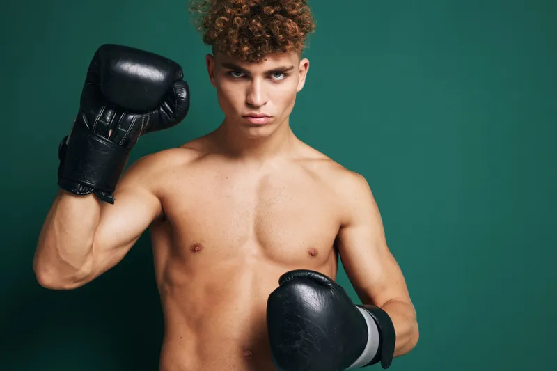 Young man with curly hair wearing black boxing gloves in fighting stance against green background. Shirtless athlete preparing for boxing training or competition.