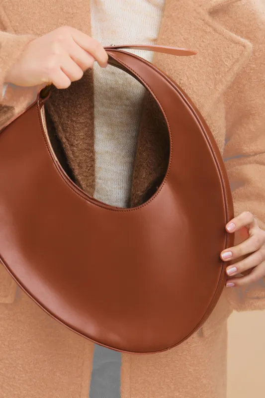 asian woman holds a circular brown handbag in a stylish pose, showcased against a neutral backdrop. warm leather, soft texture, and contemporary silhouette create a chic urban accessory moment.