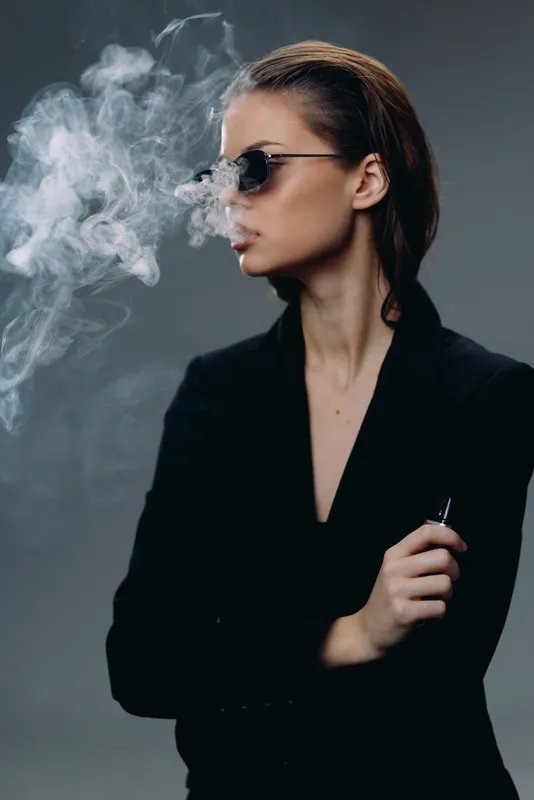 Stylish woman smoking vape with sunglasses and black outfit exhaling thick white smoke in dark studio.