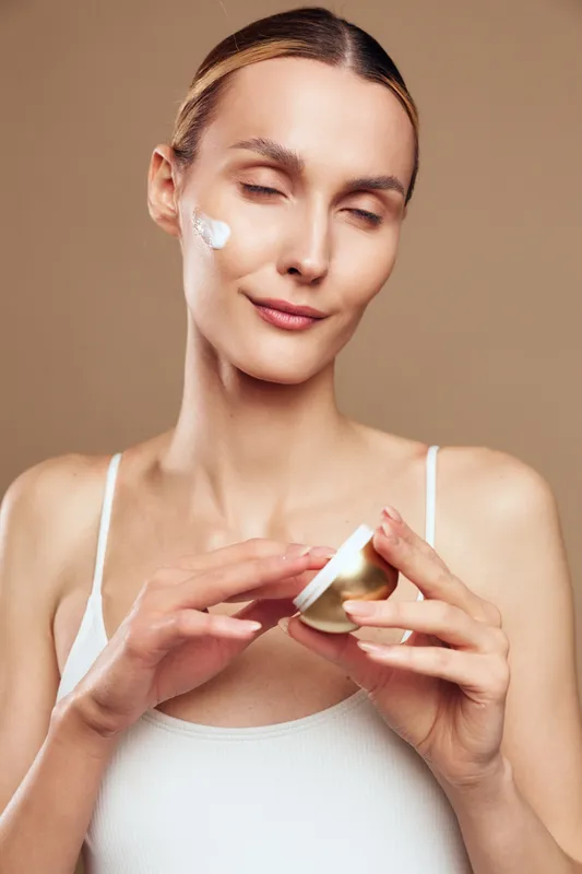 A young woman in a white tank top applies cream on her face with closed eyes. The neutral background emphasizes skincare routine and self-care concept for healthy skin.