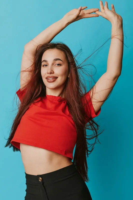 Young woman in red crop top with long hair posing with raised arms against blue background, smiling and looking confident in casual fashion style portrait.