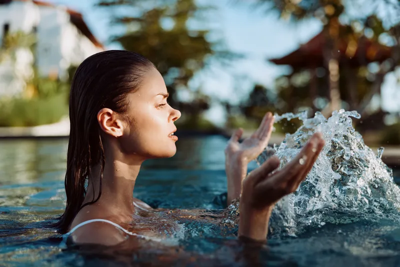 A young girl in a pool splashes water with closed eyes under bright sun, capturing a carefree lifestyle moment of outdoor fun, relaxation, and summer refreshment.