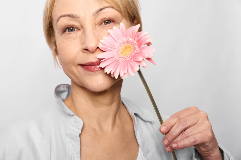 Smiling woman closeup holding a pink flower by her cheek, soft indoor light, minimal white background, relaxed portrait, gentle mood, beauty care, fresh