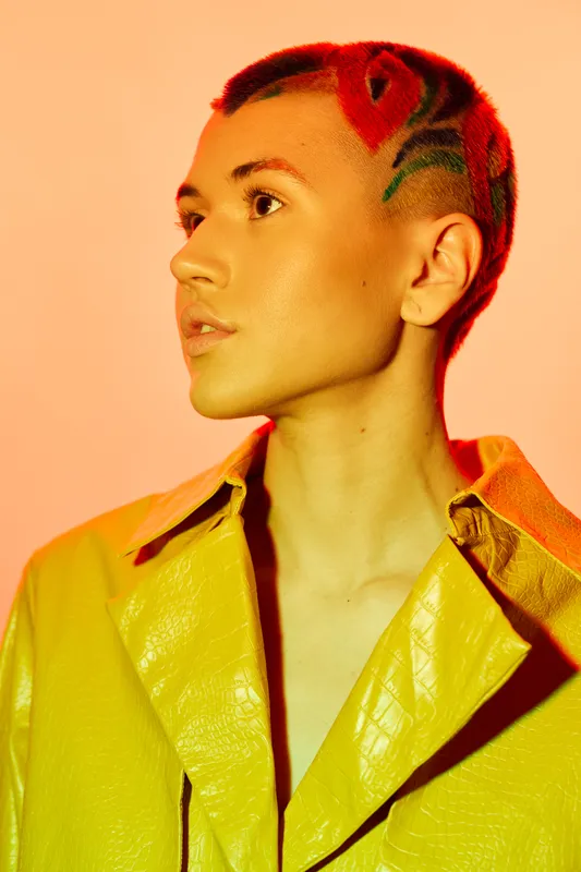 young woman with artistic red hair tattoo and yellow leather jacket looking sideways with thoughtful expression, warm orange background, creative fashion style