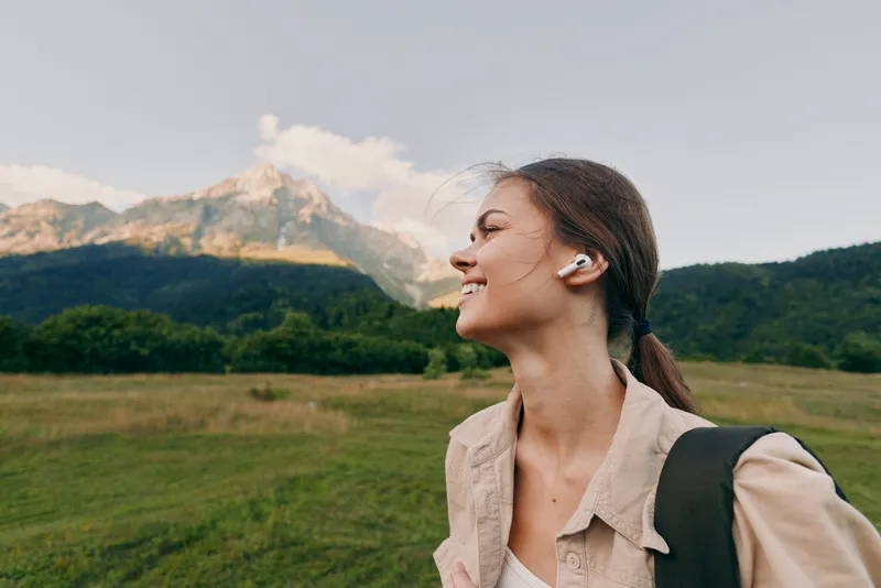Young woman with earbuds smiles enjoying a peaceful moment in a green meadow with mountains in the background under a clear sky.