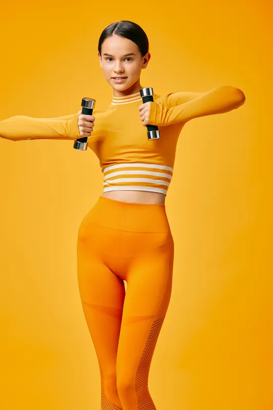 Female athlete in orange activewear lifts light dumbbells against a vivid orange studio backdrop, showcasing fitness routine, strength, and athletic confidence.