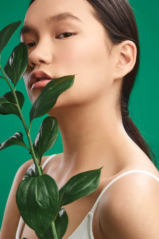 asian woman gazes softly at the camera while holding a leafy plant, set against a vivid green backdrop, creating a calm, fresh studio portrait with natural light and contemporary style.