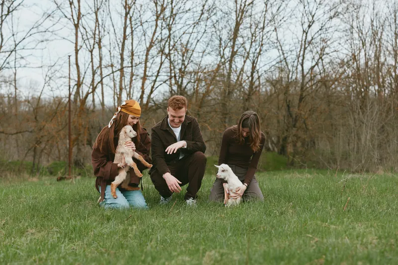 Three young adults spend time outdoors in a grassy field with two baby goats during early spring, enjoying a casual lifestyle moment together with relaxed clothing and natural surroundings.