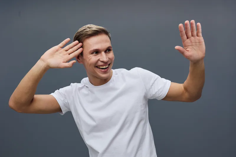Smiling man in casual white T-shirt facing camera, raising hands in friendly greeting gesture, isolated on plain gray background, studio portrait, positive emotion, people lifestyle concept
